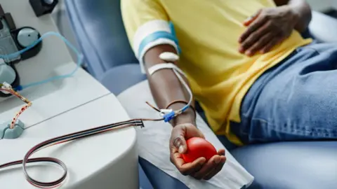 Getty Images A man wearing a yellow shirt lies on a couch giving blood with a tube attached to his arm, while holding a soft red ball in his hand 