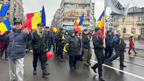 BBC A crowd of people carrying Romanian flag march through Bucharest. Most are wearing dark clothing and hats.