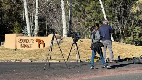 Reuters Reporters gathered outside the gated community where Hackman and his wife died. Two reporters wear denim jean pants, and stand near tripods holding up cameras. They face a sign that says 'Santa Fe Summit'