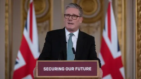 PA Media Sir Keir Starmer wearing a black suit and white shirt with green tie, standing in front of two Union Jack flags and behind a podium with the slogan 