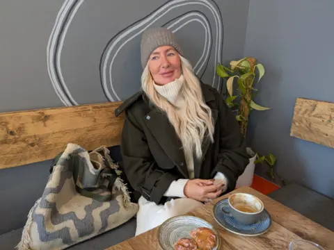 Karen Pearson sits in front of a pastry and coffee in an independent coffee shop, wearing a woolly hat and jacket