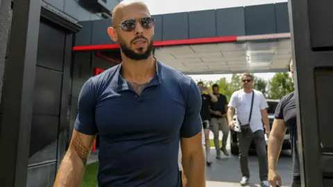 Reuters Andrew Tate, who has a bald head, black beard and is wearing sunglasses and a blue polo-neck t-shirt walks towards the camera.