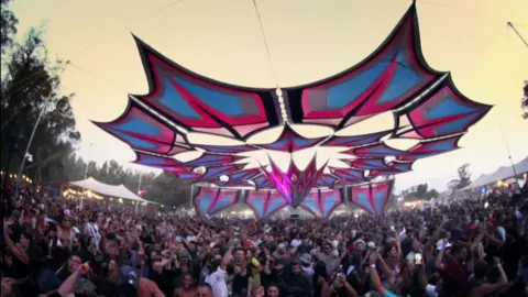 A still from footage at the Nova festival before the attack, with a large crowd dancing under a star-shaped awning in psychedelic tones of blue and pink. Some lights are on and the sky has a touch of early morning glow