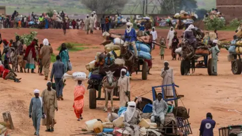 Reuters A group of people walking down a pathway in Darfur, some carrying items on their head or in their hands. A group of people sit on several different carts, led by donkeys, with items piled on the cart.