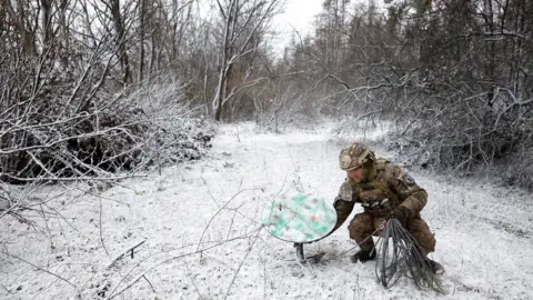 Reuters Ukrainian soldier crouches to disconnect Starlink receiver in snow-covered forest clearing on the frontline in January 2023