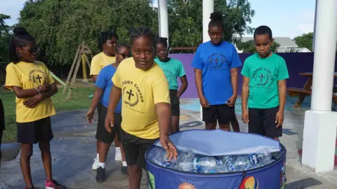 Gemma Handy A group of children in colourful T-shirts bearing the school's logo watch as one of them puts a plastic bottle into a recycling bin