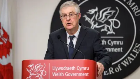 Getty Images Mark Drakeford standing at a Welsh government branded lecturn hosting a news conference during the pandemic. He has a large circular grey bilingual sign made of slate behind him saying Welsh government and first minister of Wales and a Welsh flag to his right