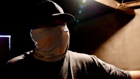 Darren Conway/BBC A man wearing a baseball cap and with a bandana covering his face, looking towards the camera as he stands in a bare, starkly lit room, much of which is in deep shadow.