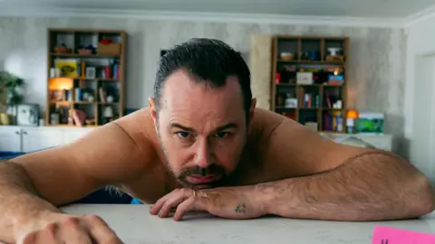 TRUE BRIT Danny Dyer is slumped on a white table, chin on his elbow and looking up at the camera. He has a dark beard and moustache and wears a gold wedding ring and no shirt. There is a small tattoo on the side of his hand. In the background are two bookshelves, one either side of him. His expression suggests he is feeling rough.