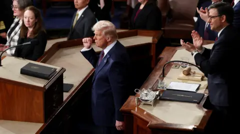 Reuters President trump is seen in an aerial view in the House chamber with his fist in the air. He is wearing a blue suit and Mike Johnson stands above him applauding.