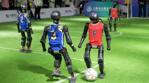 BBC/Joyce Liu Black robots in blue and red jerseys play football on a green carpet of artificial grass.