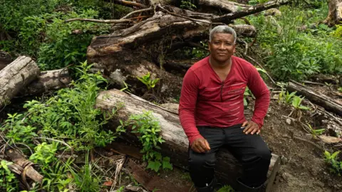 BBC / Paulo Koba Claudio Verequete sits on a felled tree, wearing a red jumper. He has short grey hair and is looking at the camera