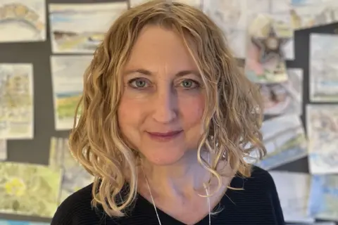 Prof Sian Bayne who has shoulder length, blonde, wavy hair. She is wearing a black jumper and a necklace. She is smiling at the camera and there is student artwork on the wall behind her.