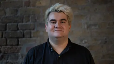 Jake Hurfurt Jake Hurfurt looks directly at the camera. He has died blond hair with black roots showing, is clean shaven and wears an open collar black shirt. Behind him is an exposed brick wall.