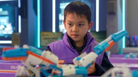 BBC/ Joyce Liu A child in a purple jacket operates a blue and orange AI-powered toy he built using code 