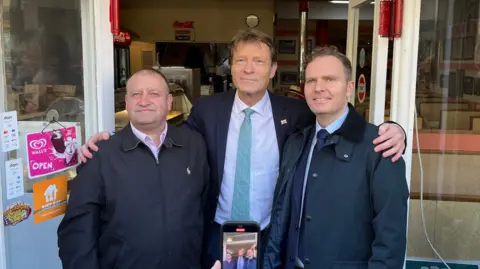 Richard Tice poses with his arms around to other men. He is wearing a dark suit, white shirt and blue tie. The man to his left is wearing a navy Ralph Lauren jacket and a pink shirt. The man to his right is wearing a navy jacket and a navy tie.