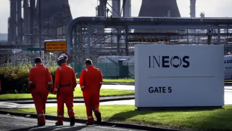 David Cheskin/PA Wire Three workers in orange boiler suits walk past a white Ineos sign. The Grangemouth plant can be seen in the background.