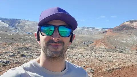 Dave Gold Dave Gold is wearing reflective sunglasses and a purple hat with a mountainous area in the background