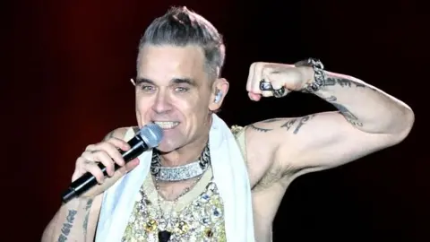 Getty Images Robbie Williams sings into a microphone on stage. He smiles wearing a sparkly vest