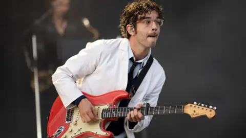 PA Media The 1975's Matty Healy, dressed in a white suit, pulls a face while playing guitar on stage at a festival in Scotland, 2024