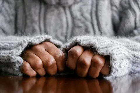 Getty Images A stock image of hands in fists, resting on a wooden surface. The person is wearing a grey woolly jumper and its sleeves are pulled up close to the person's knuckles. 