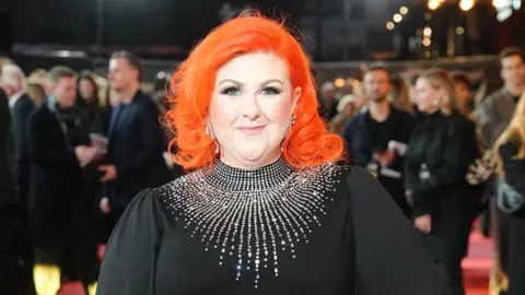 PA Media Michelle McManus smiles at the camera while on the red carpet. You can see lots of people in dark outfits blurred in the background behind her. The head and shoulders shot shows her orange, shoulder-length hair and black dress bedazzled with silver gemstones around her neck.