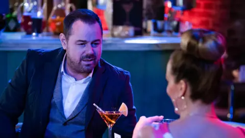 Danny Dyer looks across the table at a woman in front of a bar. He wears a grey waistcoat and a dark suit jacket and white shirt. You can see the back of her head - a blonde bun, hoop earring and pink top. She looks like Linda, his wife in EastEnders, and the setting looks like one of the bars on the EastEnders set. There is an orange drink in a cocktail class with a straw between them. On the bar are pitchers of different coloured drinks. The woman looks like she is smiling but you cannot see much of her face. He is looking into her eyes.