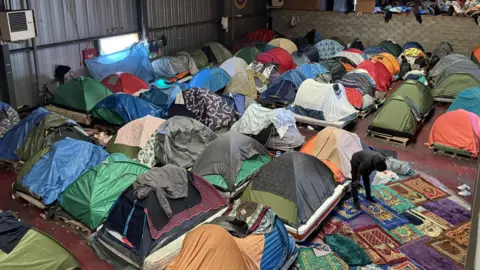 Michael Keohan/BBC Several tents inside a warehouse. Prayer mats form a makeshift area for those seeking to pray. 