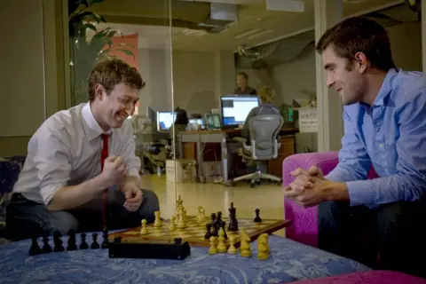 Eyevine Two people - one of them Mark Zuckerberg - are playing chess. The chess board is perched on a blue, patterned foot stool. Mark Zuckerberg is playing with white pieces, his opponent black. Mr Zuckerberg is clenching his right fist and smiling.