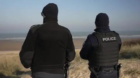 Two French police officers patrol the beach at Sangatte. They are both facing away from the camera. 