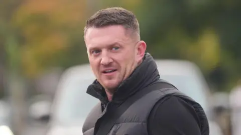 PA Media Tommy Robinson looking to the right front of the photo. He is wearing a black top, a black gilet, with a bag strap across one shoulder. He has light stubble.