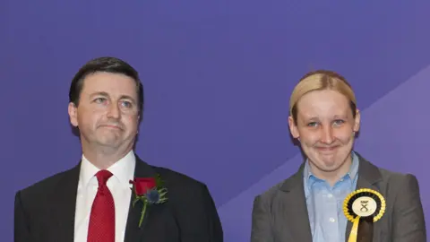 Getty Images The election count in 2015 - Douglas Alexander -  a dark haired man with a suit, red tie and red rosette, stands next to Mhairi Black, who has  blonde hair and is wearing a grey suit and an SNP rosette