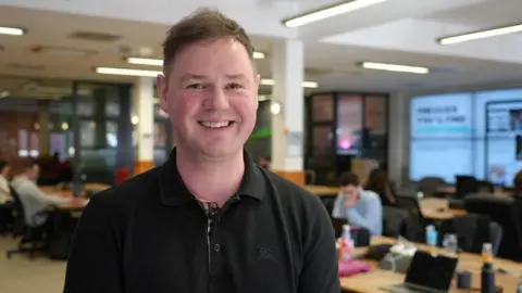 A portrait shot of James Robinson, a man in a black polo shit with short brown hair. A blurred office behind him contains people at desks with open laptops.