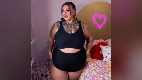 Sophie Scott Sophie who is a UK size 24 to 26 model wearing a black sports bra and cycling shorts and glamorous make up, posing in a pink bedroom in an Instagram-style shot