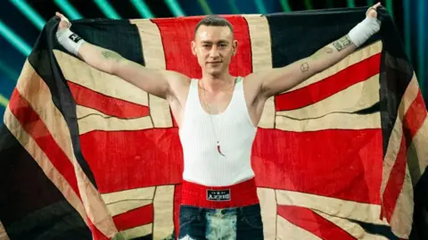 Getty Images Olly Alexander at Eurovision in 2024. Olly holds the Union Jack flag over his shoulders while wearing a white tank top and red boxer shorts visible over his denim jeans