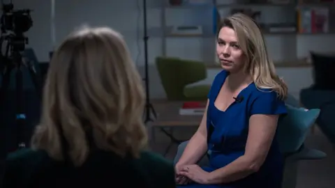 Emma Lynch / BBC Sarah Wynn-Williams - a woman with long blond hair, wearing a blue dress - sits on a chair opposite the BBC's Katie Razzall, who has her back to the camera.