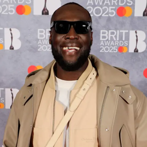 Getty Images Stormzy smiles for the camera while wearing sunglasses, as he poses on the Brit Awards red carpet.