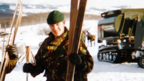 Stuart Anderson A solider wearing a camouflage jacket and green winter hat, holding skis on a snowy mountain with a military vehicle on tracks behind him.