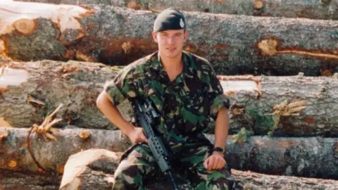 Stuart Anderson Soldier wearing green beret, camouflage shirt and trousers, with his sleeves rolled up, holding a machine gun, sat on a pile of very large logs.