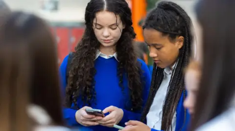 Getty Images Stock image of two female schoolchildren looking at their phones while in school