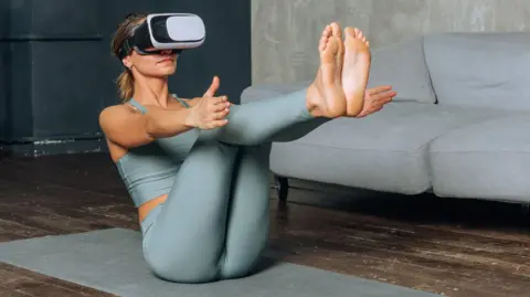 Getty Images A woman strikes a yoga pose on a mat in a living room while wearing a VR headset.