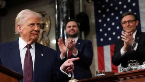 Reuters Donald Trump looks to his left, pointing with his index finger as JD Vance and Speaker of the House Mike Johnson stand behind him clapping their hands