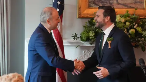 PA Media JD Vance and Micheál Martin shaking hands in front of the US flag. They are both wearing suits and smiling.