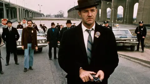 Getty Images Policeman Gene Hackman is shown in a movie still walking toward the camera. He is holding a gun in his hands with policemen and police vehicles in the background