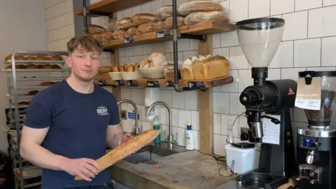 Toby Johnstone, manager of Flour, Water, Salt, holding a loaf of bread in front of shelves of bread in the cafe