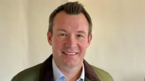 BBC Man with dark brown hair posing for a photo with a cream wall behind him, wearing a pale blue shirt and green jacket with brown collar.