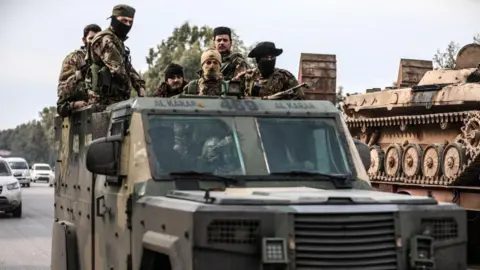 Getty Images Armoured vehicle with six soldiers standing in the back of it. Four are wearing hats and three are wearing face coverings. The vehicle is on a road and in the background there is a tank.