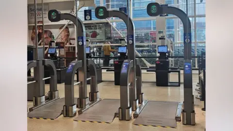 Reddit Three giant scales in front of Tesco Scan as you Shop checkouts. They have a large metal plate on the ground and barriers either side with numbers one to three above. Reddit users have posted that they look like airport security scanners.