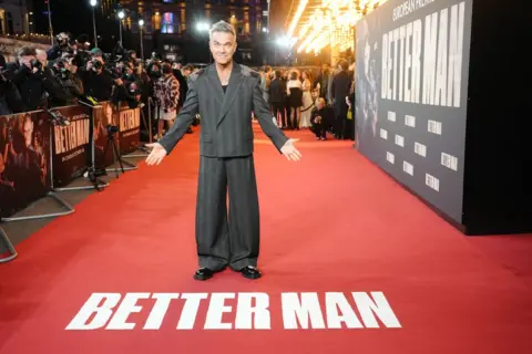 PA Media Robbie Williams wears a dark striped, slightly baggy suit on the red carpet for his film Better Man. Photographers can be seen behind the barrier in the background. Robbie gestures towards the red carpet in front of him which has the words 