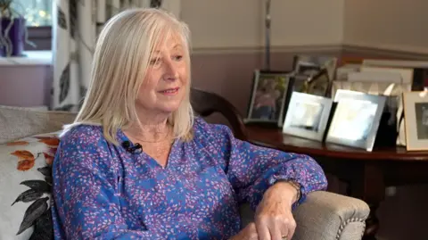 Linda Kelly sits in an armchair in her home, wearing a purple blouse, with framed photos behind her on a table